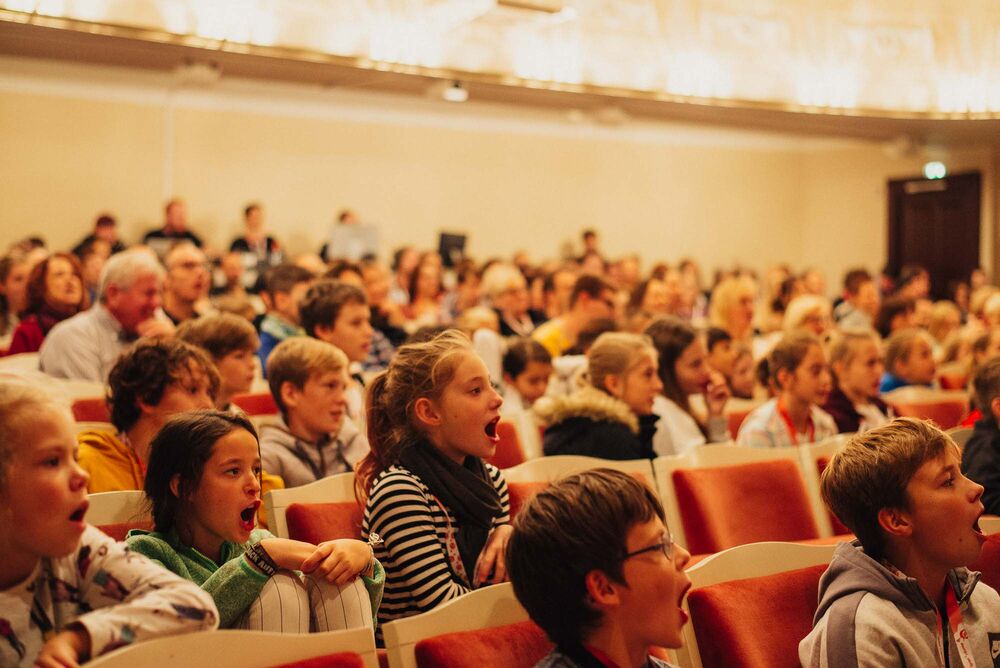 Erwachsene und viele Kinder in einem Theatersaal. Einige Kinder rufen etwas mit weit offenem Mund.